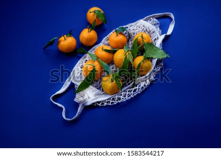 Tangerines (mandarines) with young green leaves in a textile 
reusable fabric mesh  on a deep phantom blue background. Zero waste concept. Close up. Copy space. Flatlay. 