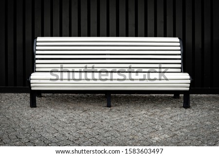 A white bench on the cobblestones against a dark vertical ribbed metal fence