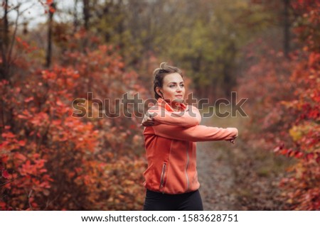 Young attractive sport woman in sportswear doing stretching exercise for hand in autumn forest with reddened leaves of trees. Warm up before training. Healthy lifestyle concept