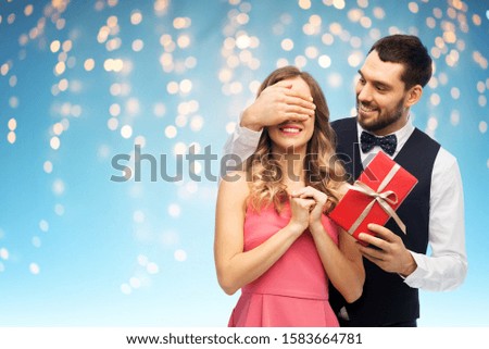 valentines day, couple, relationships and people concept - happy man giving woman surprise present over holiday lights on blue background