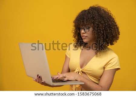 Portrait of a smiling young curly hair mixed race girl holding laptop computer isolated over yellow background.