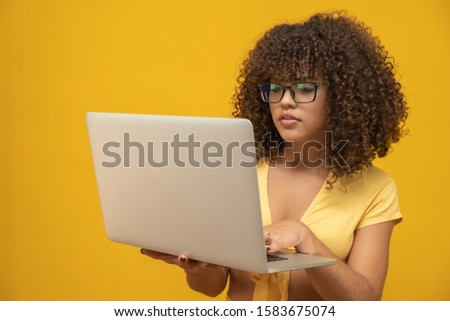 Portrait of a smiling young curly hair mixed race girl holding laptop computer isolated over yellow background.