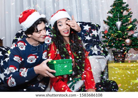 Romantic multiethnic couple exchanging gifts near decorated christmas tree. Smiling man in sweater giving christmas present surprise to chinese woman.
