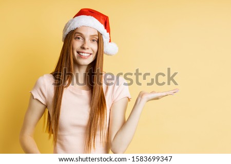 Surprised lovely caucasian ginger girl in Santa hat and t shirt, holding copy space, posing with open palm, showing or presenting your product, item isolated on yellow background. Advertising concept.