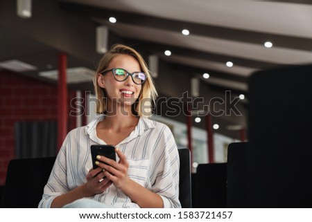 Photo of smiling blonde woman wearing eyeglasses typing on smartphone while sitting in conference hall