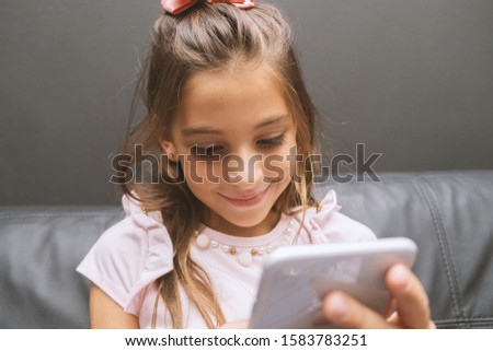 Beautiful little girl playing game or watching video on smartphone mobile. Girl watching cartoons or browsing internet, copy space. Portrait of little girl using smartphone while sitting.
