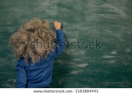 Cheerful smiling little kid (boy) against green chalkboard. Looking at blackboard. School concept