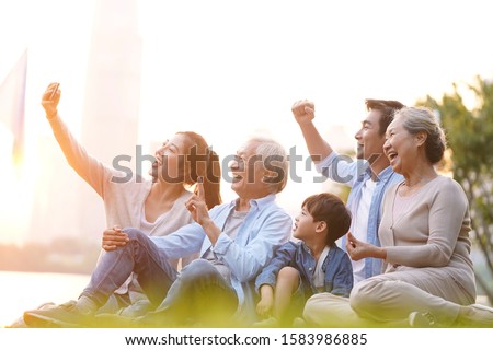 three generation happy asian family sitting on grass taking a selfie using mobile phone outdoors in park