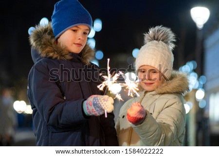 Two cute young children, boy and girl in warm winter clothing holding burning sparkler fireworks on dark night outdoors bokeh background. New Year and Christmas celebration concept.