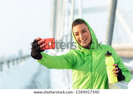 Jogging woman taking selfie by mobile phone with bridge on snow on background