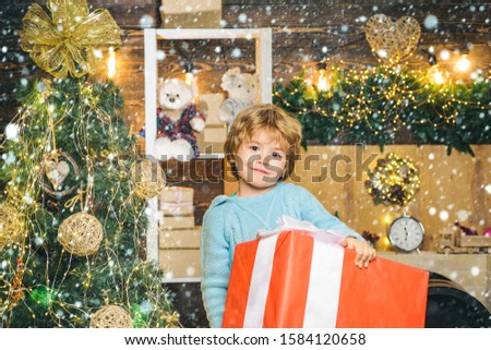 Happy child holding a giant red gift box with both hands. Christmas Celebration holiday. Happy little child dressed in winter clothing think about Santa near Christmas tree