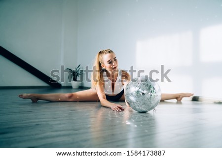 Athletic slim woman with blonde long hair dressed in black and white bodysuit doing splits on floor before mirror disco ball in studio