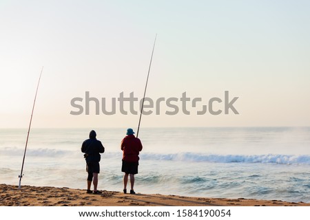 Fishermen fishing  on beach shoreline morning silhouetted facing horizon winter mist over waves and sea water holding long surf rods rear landscape.