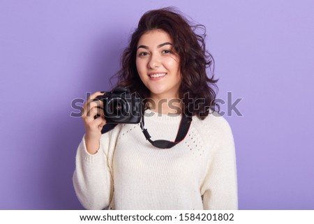 Picture of charming magnetic young female with curly black hair, holding photo camera, taking photos, smiling sincerely, wearing white sweater, spending time alone, getting pleasure. Hobby concept.
