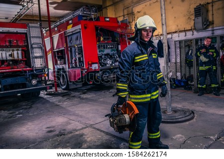 A fireman with uniform and helmet holding a chainsaw with fire truck in the background
