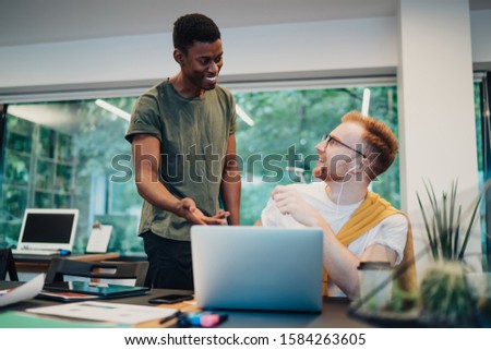 Smiling smart diverse young coworkers in casual apparel having discussion surfing on laptop in office space opposite of big window  