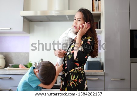 Busy and stressed mother talking on phone with one hand carrying her little infant baby and bother boy crying and annoying on kitchen background. Housewife overwork Royalty-Free Stock Photo #1584282157