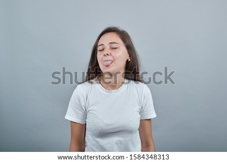 Teen in white t-shirt shows her tongue in jest. Gils looks childish and windy. Behind her is gray wall. Hair is flowing down.