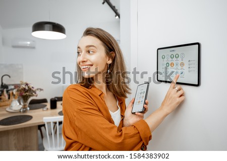 Young woman controlling home with a digital touch screen panel installed on the wall in the living room. Concept of a smart home and mobile application for managing smart devices at home Royalty-Free Stock Photo #1584383902