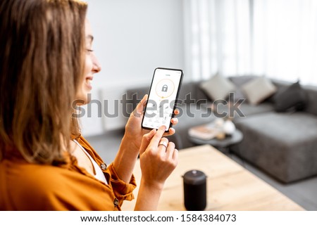 Young woman holding smart phone with launched security application at home. Concept of controlling and managing home security from a mobile device Royalty-Free Stock Photo #1584384073