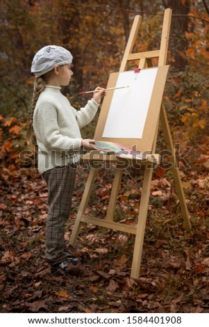 Beautiful girl artist with paints and palette, paints a picture on an easel in the autumn forest. Creative work in nature and fresh air. Useful creativity and relaxation for children.