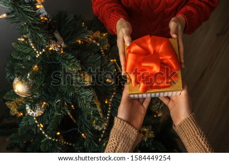 Hands of man giving a Christmas gift with red bow to woman near the christmas tree.