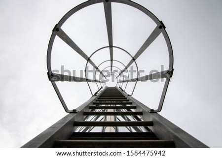 Architectural photo of metal service ladder with safety cage against bright white sky background. Worm's eye view. 