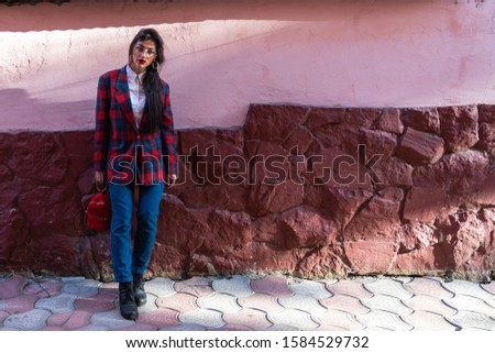 Street sunny photo of the beautiful stylish young woman in glasses, dressed in blue jeans and plaid jacket, holds little red backpack in her hand and looking at the camera, pink and red wall on the