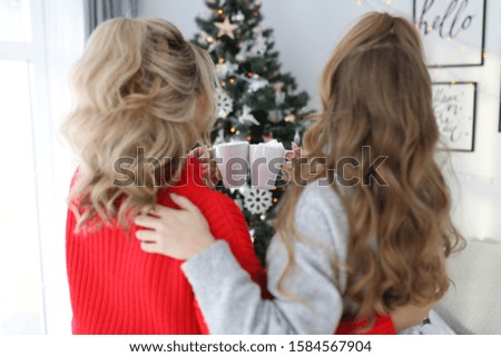 Two cute beautiful girls girlfriends sisters lovers in cozy bright red sweaters laugh smile and pose against the background of a Christmas tree in a decorated beautiful bright room.