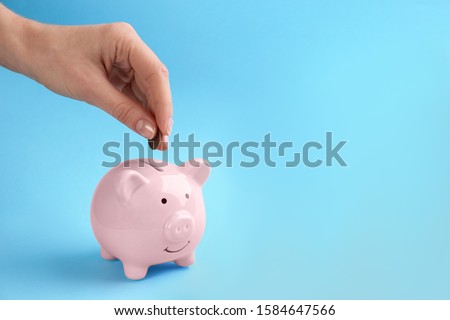 Woman putting coin into piggy bank on light blue background, closeup. Space for text Royalty-Free Stock Photo #1584647566