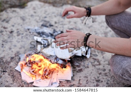 Young man, with occult hippie bracelets on his hands, burning bonfire of paper on abandoned construction area industrial zone in summer. Close-up picture of hands, burning fire from paper.