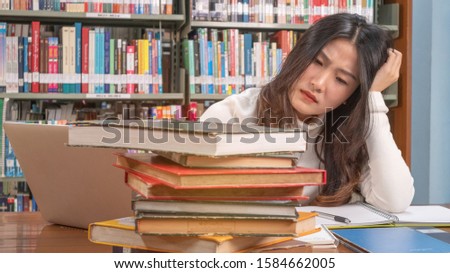 Close up young asian woman sitting and thinking seriously while looking stack of book on desk in library with bookshelf background prepare exam university or school, education and searching concept.