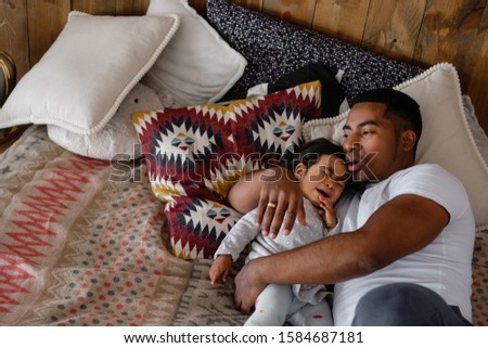 Kind young African American father calms his little mixed race crying two-year-old daughter lying on the bed in a cozy room. The concept of a caring father