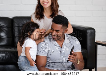 Surprised young funny african-american father shows his charming little daughter cartoon using smartphone and high speed internet and sitting on floor amid unidentified caucasian mom sitting on a sofa