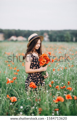 
Beautiful young brunette model girl in a hat walks in a flower field with a bouquet of poppies. Fragrant field of red flowers.