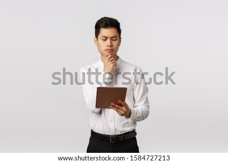 Pensive attractive asian businessman making hard choice, looking at digital tablet and thinking what order girlfriend online, reading documents on device screen, frowning thoughtful, white background