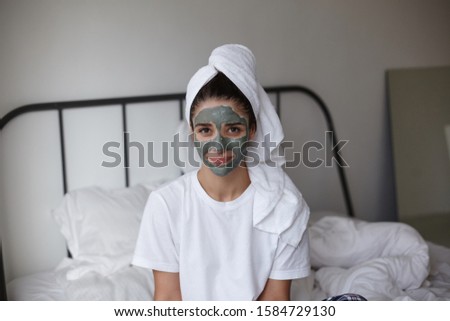 Portrait of lovely brown-eyed brunette woman wearing white towel on her head while keeping mask on her face, looking positively to camera and smiling slightly, isolated over home interior