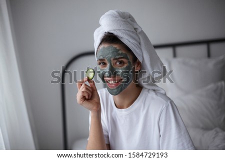 Positive attractive young dark haired woman with cosmetic mask of blue clay on her face applying fresh cucumber on her eye and smiling cheerfully to camera, isolated over home interior