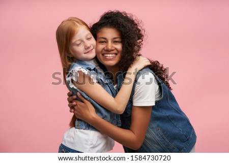 Cheerful attractive young dark skinned brunette woman with long curly hair smiling happily to camera and cuddling cute positive redhead female kid, isolated over pink background
