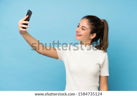 Young brunette girl over isolated blue background taking a selfie with the mobile