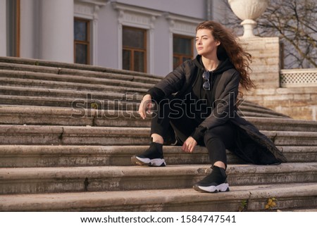 Brooding girl holding chin looks at blank space. Beautiful woman with green eyes, long curly dark hair is wearing black jacket, sweater, jeans, sneakers, sitting on steps of stairs, staring away