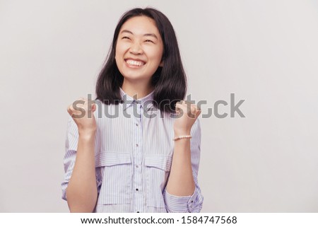 Winner concept. Girl excited for success with arms raised celebrating victory smiling. Businesslike beautiful young woman of Asian appearance dressed in striped office shirt stands isolated background