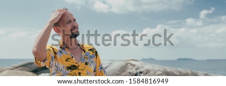 A man stands against a tropical landscape. Young guy wearing a hawaiian shirt on a tropical island. Handsome man and bright positive emotions. Banner