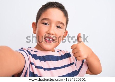 Beautiful kid boy wearing casual striped t-shirt make selfie over isolated white background happy with big smile doing ok sign, thumb up with fingers, excellent sign