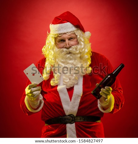 Santa Claus holds playing cards and a bottle with alcohol in his hands, drinks and poses on a dark red background