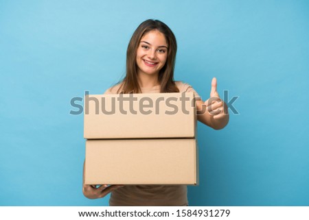 Young brunette girl over isolated blue background holding a box to move it to another site with thumb up