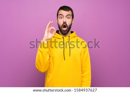 Handsome man with yellow sweatshirt surprised and showing ok sign