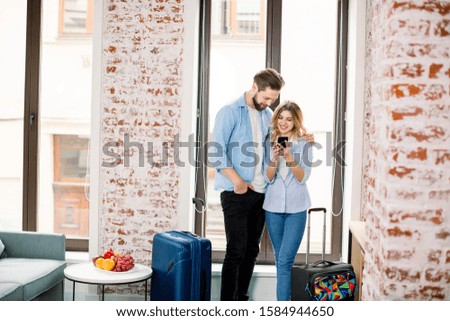 Happy couple looking for the photos in a smart phone standing over the window background in modern hotel room with brick walls