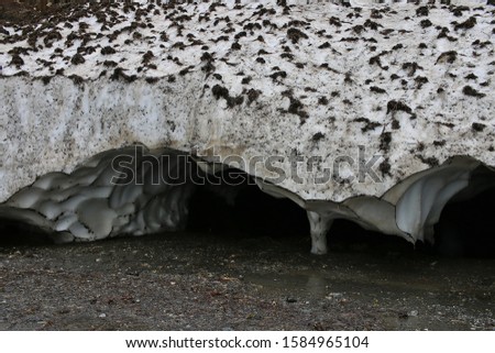 Pure snow caverns and hollows beneath very thick melting snow layers depicting the formation of ice crevasses on glaciers which are very perilous to cross over. The bottom is wet dirty ground