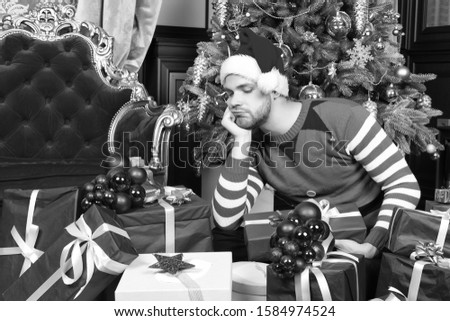 Guy in santa hat, elf costume look at present boxes with thinking face at Christmas tree. New year, xmas, eve, holidays preparation, celebration. Boxing day concept.